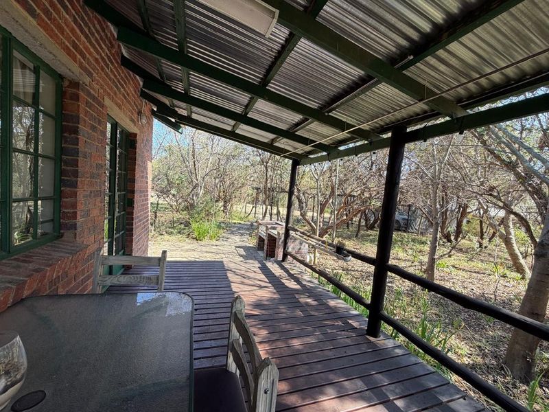 A covered outdoor patio featuring timber decking, a built-in brick braai, and a corrugated metal roof, overlooking the surrounding farm landscape.