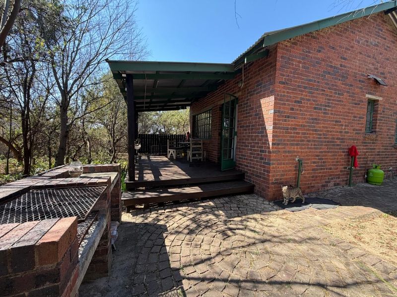 A charming outdoor entertainment area featuring a built-in brick braai, timber decking, and a covered patio space set against a rustic brick farmhouse exterior.