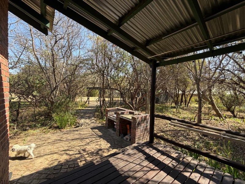 Covered patio area featuring timber decking, a built-in brick braai, and direct access to a wooded garden setting.