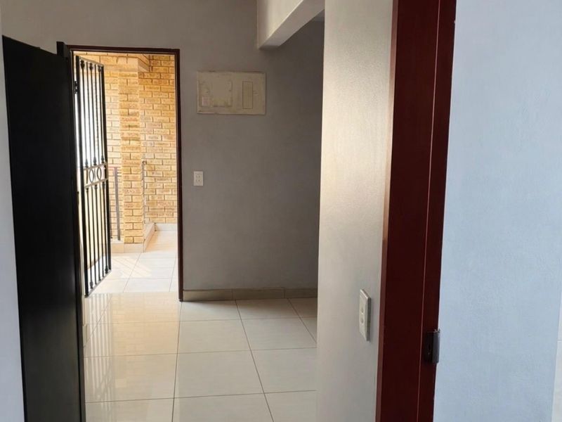 A clean, tiled hallway leading to an exterior entrance with a security gate, featuring neutral wall tones and a built-in electrical distribution board.