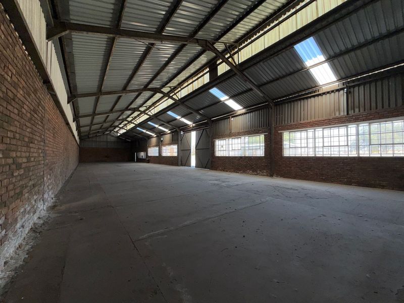 Spacious industrial warehouse featuring exposed brick walls, high corrugated metal roofing with integrated skylights, and large steel-framed windows providing ample natural light.