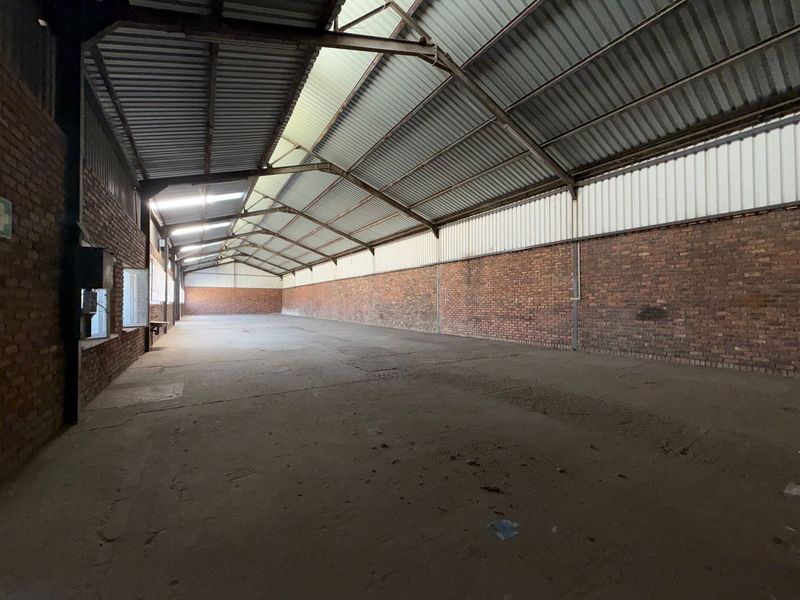 A large, open-plan industrial warehouse space featuring exposed brick walls, concrete flooring, and high-pitched corrugated metal roofing with steel trusses.