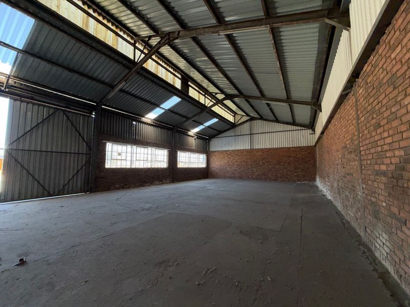 Spacious industrial warehouse unit featuring exposed brick walls, concrete flooring, high-pitched corrugated metal roof with steel trusses, and large industrial windows.