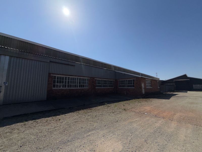 Exterior view of an industrial warehouse building featuring brick and corrugated metal construction with large steel-framed windows and a concrete yard area.