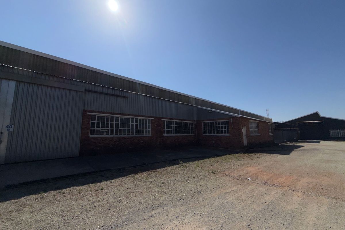 Exterior view of an industrial warehouse building featuring brick and corrugated metal construction with large steel-framed windows and a concrete yard area.