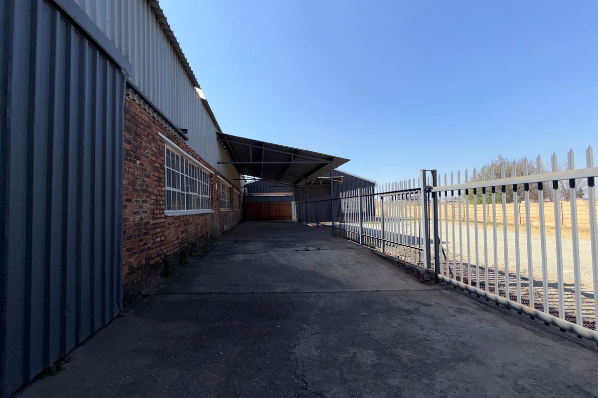 Exterior view of an industrial property featuring a concrete driveway, covered loading area, corrugated metal cladding, and secure steel palisade fencing.