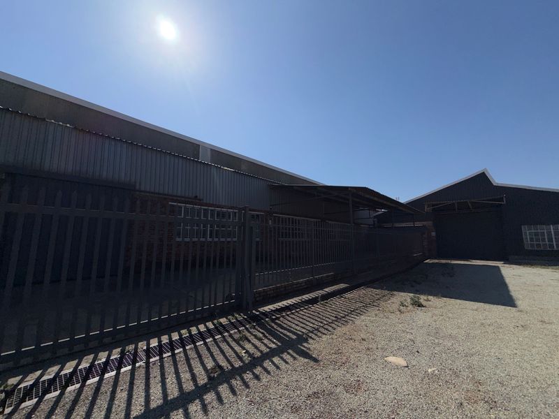 Exterior view of an industrial property featuring corrugated metal cladding, secure steel palisade fencing, and a gravel driveway area.