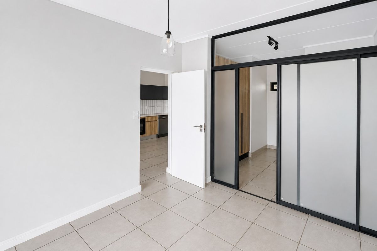 A bright, modern living area featuring neutral tiled flooring, a sleek black-framed glass partition, and contemporary lighting fixtures, leading into an adjacent kitchen space.