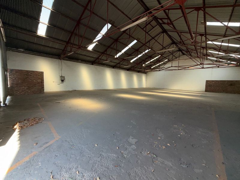 Spacious industrial warehouse featuring high exposed steel roof trusses, natural light from skylights, concrete flooring, and brick wall accents.