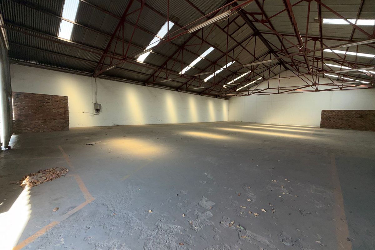 Spacious industrial warehouse featuring high exposed steel roof trusses, natural light from skylights, concrete flooring, and brick wall accents.