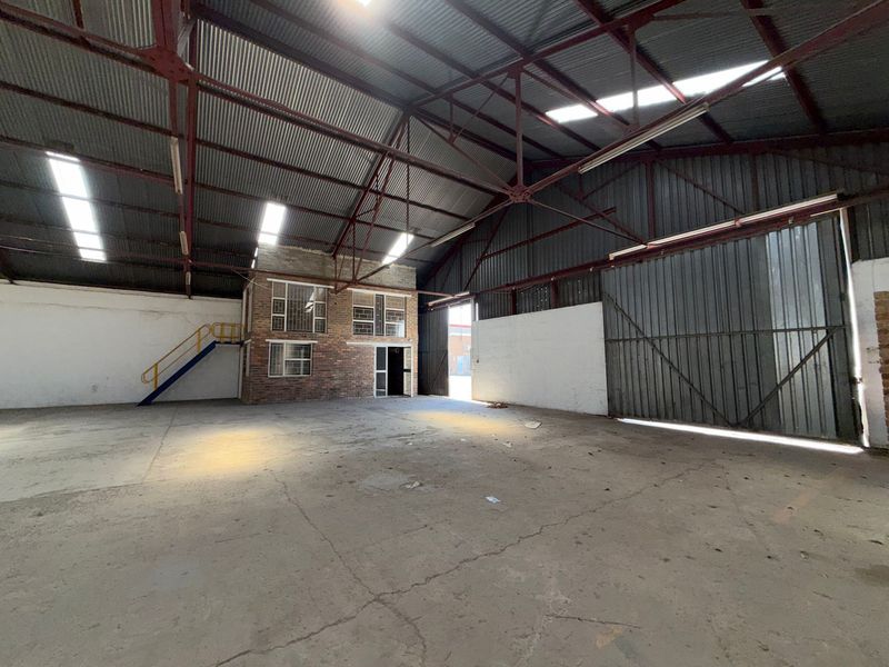Spacious industrial warehouse featuring high ceilings with exposed steel trusses, concrete flooring, a mezzanine office area, and a large sliding access door.