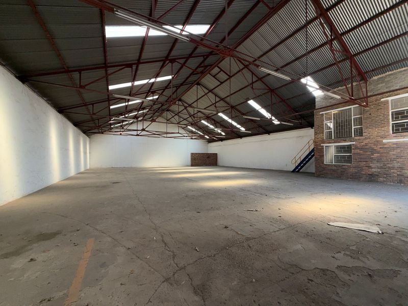 Spacious industrial warehouse featuring exposed steel roof trusses, concrete flooring, natural light from skylights, and an integrated brick office or storage section.