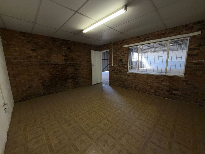An empty industrial office space featuring exposed brick walls, suspended ceiling tiles, and security bars on the window.