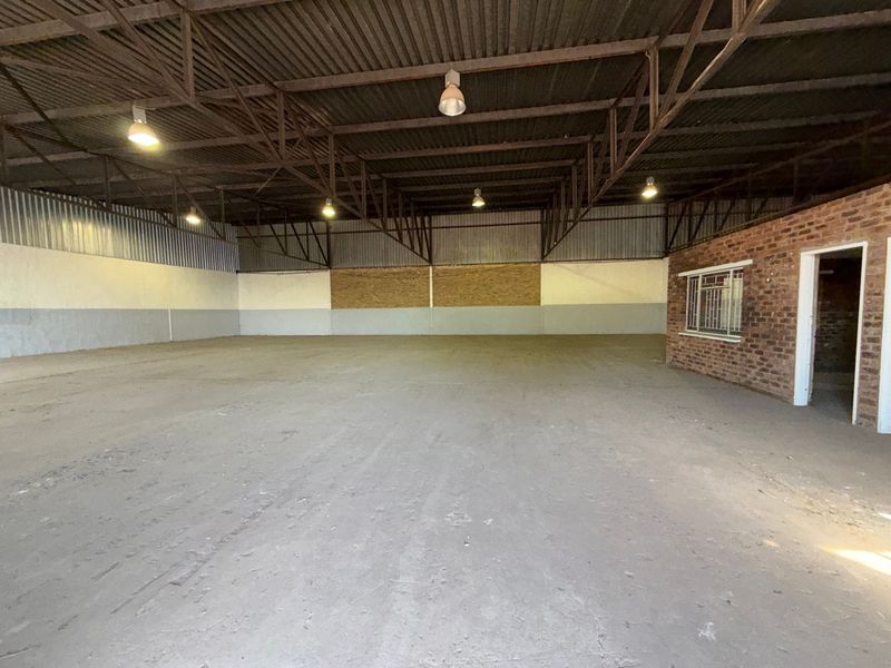 Spacious industrial warehouse unit featuring high exposed steel roof trusses, concrete flooring, and an integrated brick-walled office or utility section.