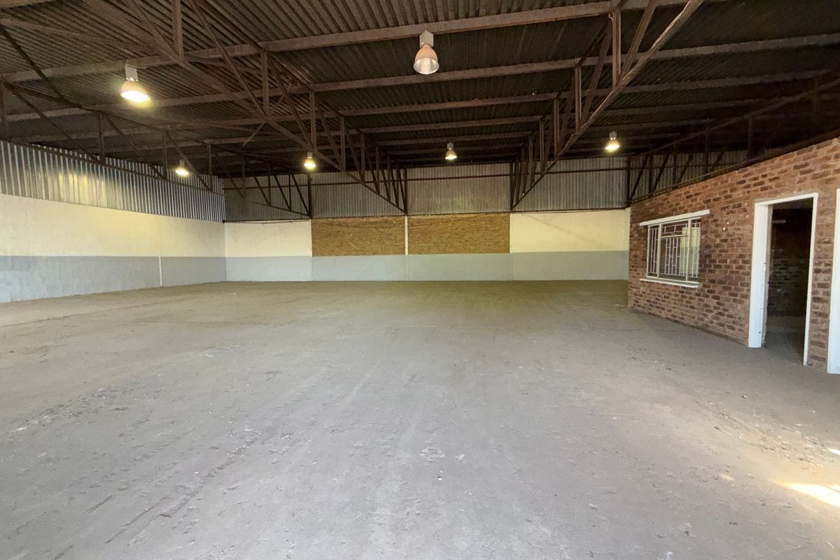 Spacious industrial warehouse unit featuring high exposed steel roof trusses, concrete flooring, and an integrated brick-walled office or utility section.