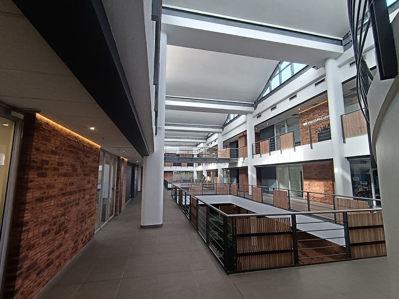 A modern, spacious office building atrium featuring exposed brick walls, tiled floors, and a multi-level design with metal railings and abundant natural light.