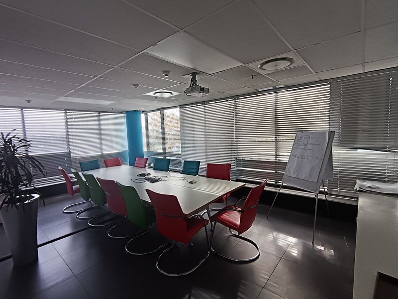 A functional office meeting room featuring a suspended ceiling, recessed lighting, ceiling-mounted projector, and large windows with venetian blinds.