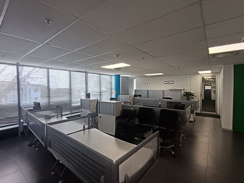 A professional open-plan office space featuring large windows with venetian blinds, suspended ceilings, recessed lighting, and modern built-in workstation partitions.