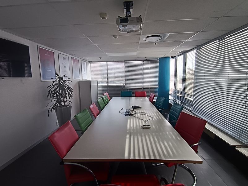 A functional office boardroom featuring a large meeting table, suspended ceiling with integrated projector, and extensive window coverage with adjustable venetian blinds.