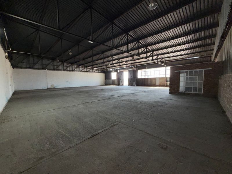Spacious industrial warehouse featuring high ceilings with exposed steel trusses, concrete flooring, and large steel-framed windows providing ample natural light.