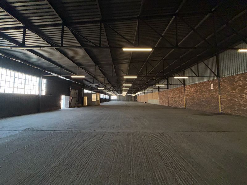 Expansive industrial warehouse space featuring high ceilings with exposed steel trusses, concrete flooring, and natural light through large steel-framed windows.