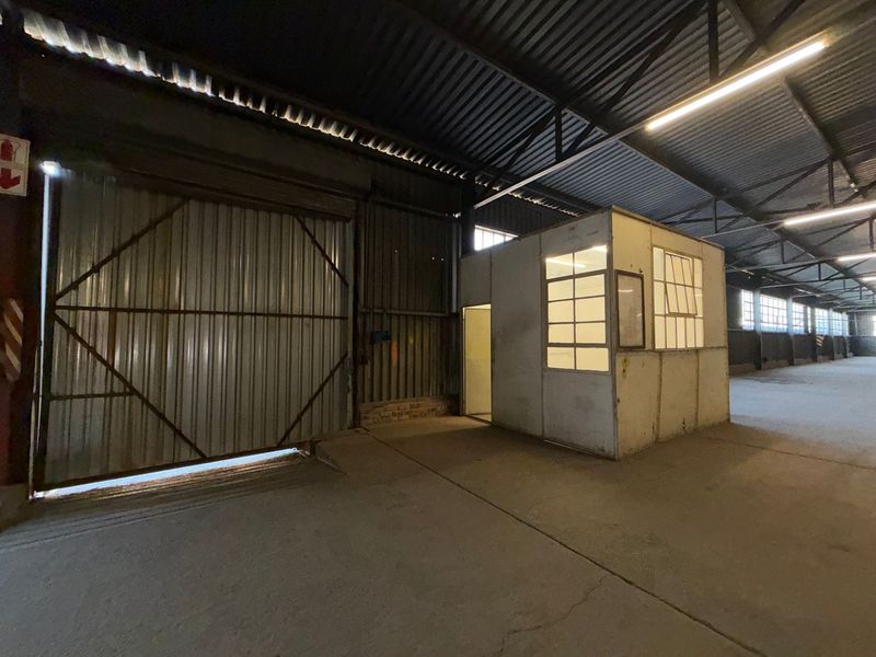 Spacious industrial warehouse area featuring high ceilings with exposed steel trusses, a large roller shutter door, concrete flooring, and a standalone internal office pod.