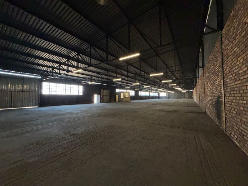A large, open-plan industrial warehouse space featuring exposed steel roof trusses, concrete flooring, and brick wall finishes with ample natural light.
