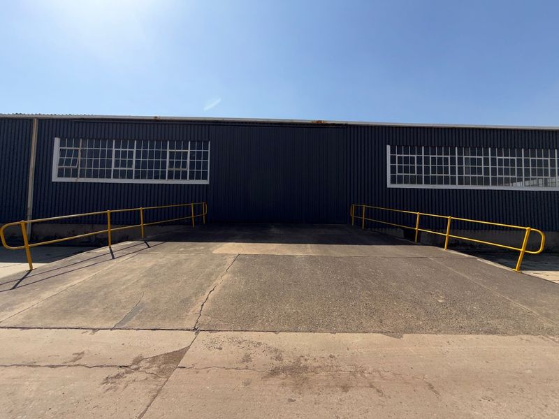 Industrial exterior view featuring a concrete loading ramp, yellow safety railings, and large sliding door access to the warehouse facility.
