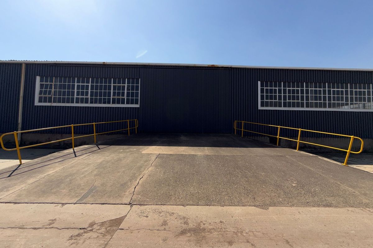 Industrial exterior view featuring a concrete loading ramp, yellow safety railings, and large sliding door access to the warehouse facility.