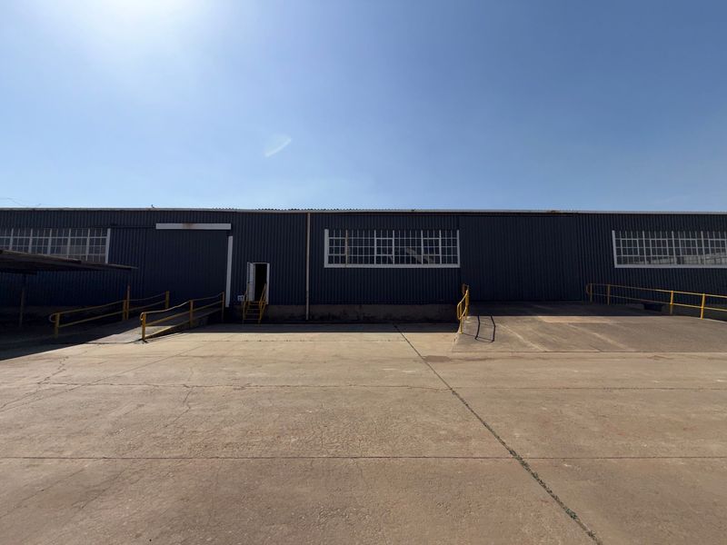 Industrial warehouse exterior featuring a large concrete loading apron, multiple loading ramps with safety railings, and corrugated metal facade with industrial windows.