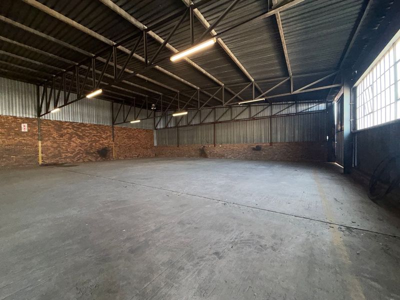 Spacious industrial warehouse unit featuring exposed steel roof trusses, durable concrete flooring, brick wall accents, and ample natural light through large steel-framed windows.