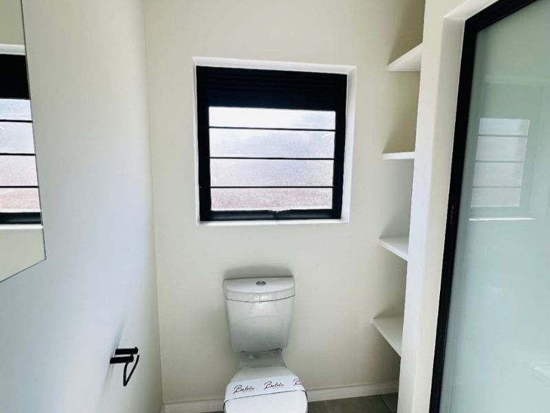 A clean, modern bathroom featuring a toilet, built-in shelving, tiled flooring, and a frosted glass shower door with a black frame.