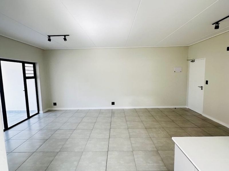 A spacious, empty open-plan living area featuring neutral tiled flooring, modern track lighting, and a sliding glass door leading to an outdoor space.