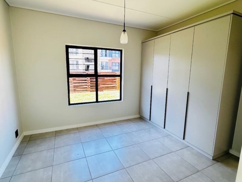 A bright, empty bedroom featuring neutral tiled flooring, built-in cupboards, and a window with security bars.