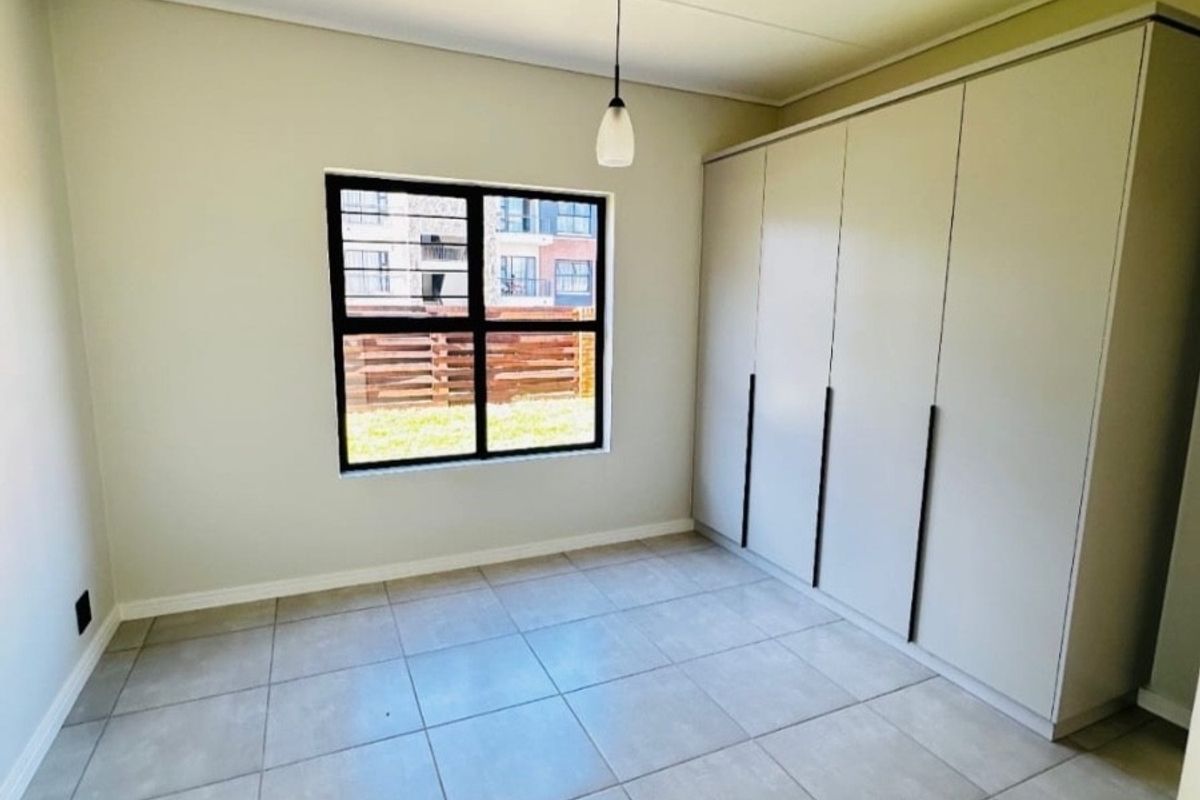 A bright, empty bedroom featuring neutral tiled flooring, built-in cupboards, and a window with security bars.