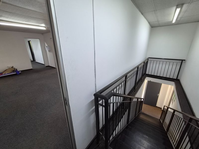 A functional industrial hallway featuring a staircase with metal balustrades, carpeted flooring, and suspended ceiling lighting, providing access to adjacent office spaces.