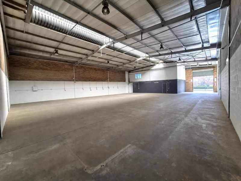 Spacious industrial warehouse unit featuring high ceilings with exposed steel trusses, concrete flooring, and a large roller shutter door for easy access.
