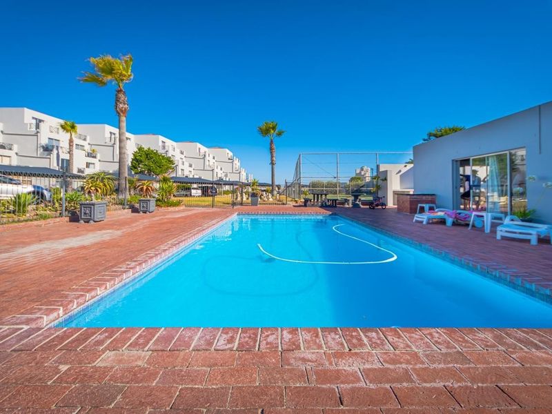 Communal swimming pool area featuring brick paving, perimeter fencing, and a built-in braai facility within an apartment complex setting.