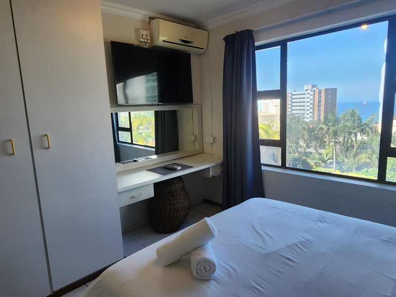 A bedroom featuring a built-in wardrobe, vanity unit, split-system air conditioner, and a large window offering scenic coastal views.