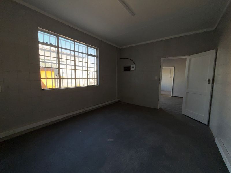 An empty industrial office space featuring concrete flooring, security bars on the window, and an exposed electrical distribution board.