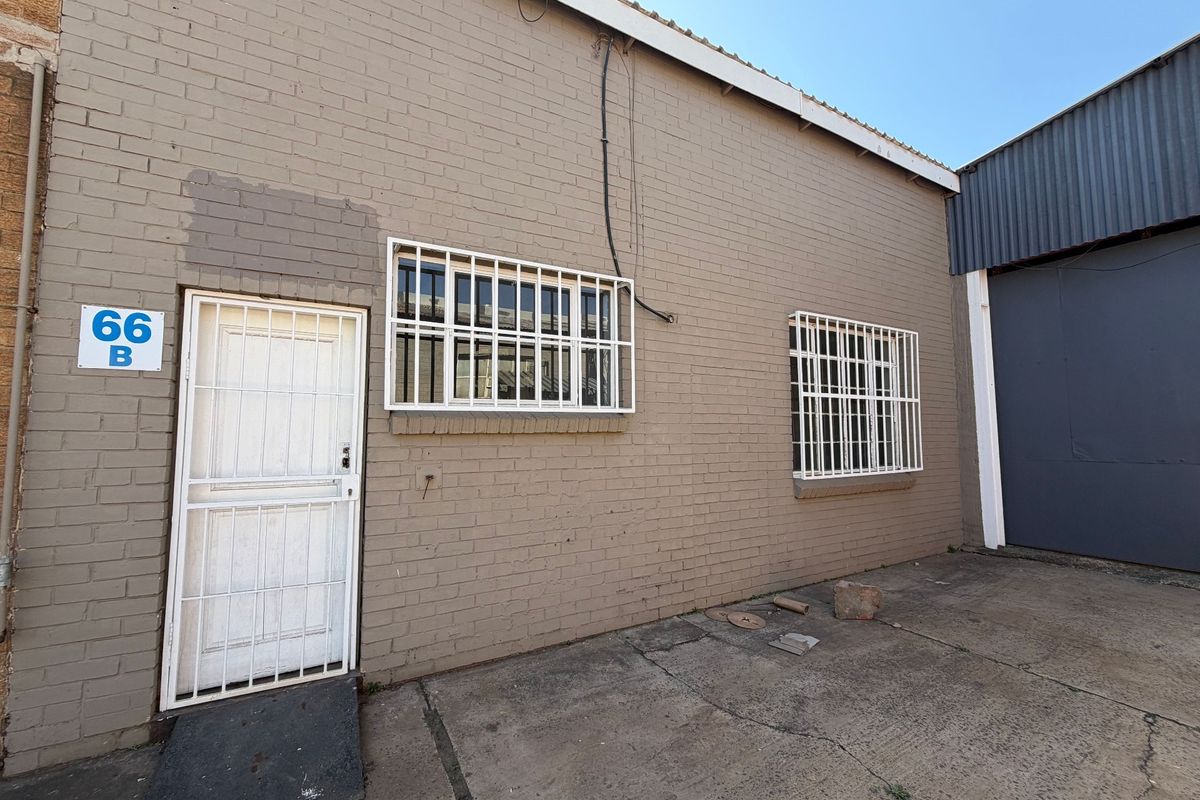 Rear exterior view of an industrial property featuring a brick facade, security-gated entrance, barred windows, and concrete paved yard area.