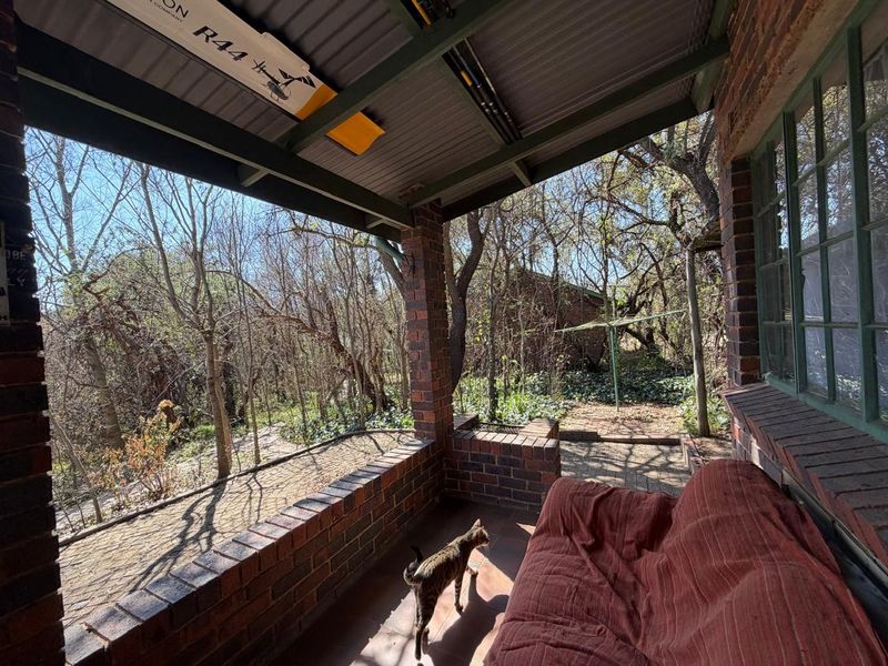 A covered brick patio area overlooking a wooded garden, featuring exposed brick pillars and a corrugated metal roof structure.