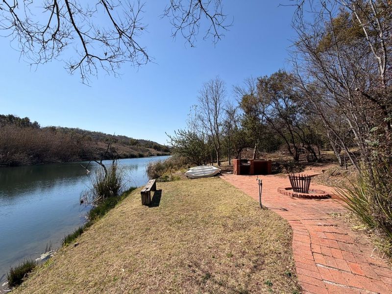 Outdoor waterfront entertainment area featuring a built-in brick braai, fire pit, and paved walkway overlooking the river.
