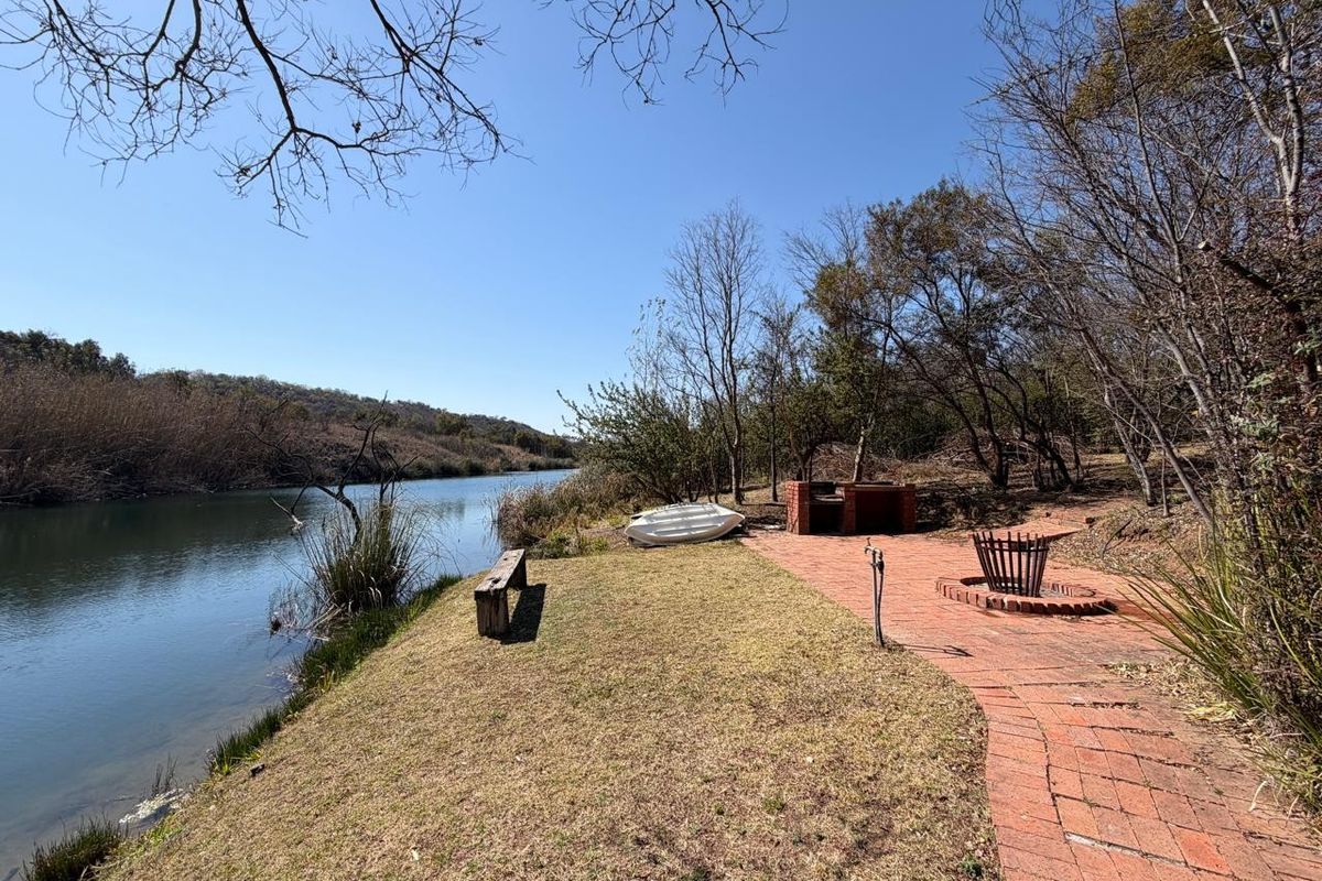 Outdoor waterfront entertainment area featuring a built-in brick braai, fire pit, and paved walkway overlooking the river.