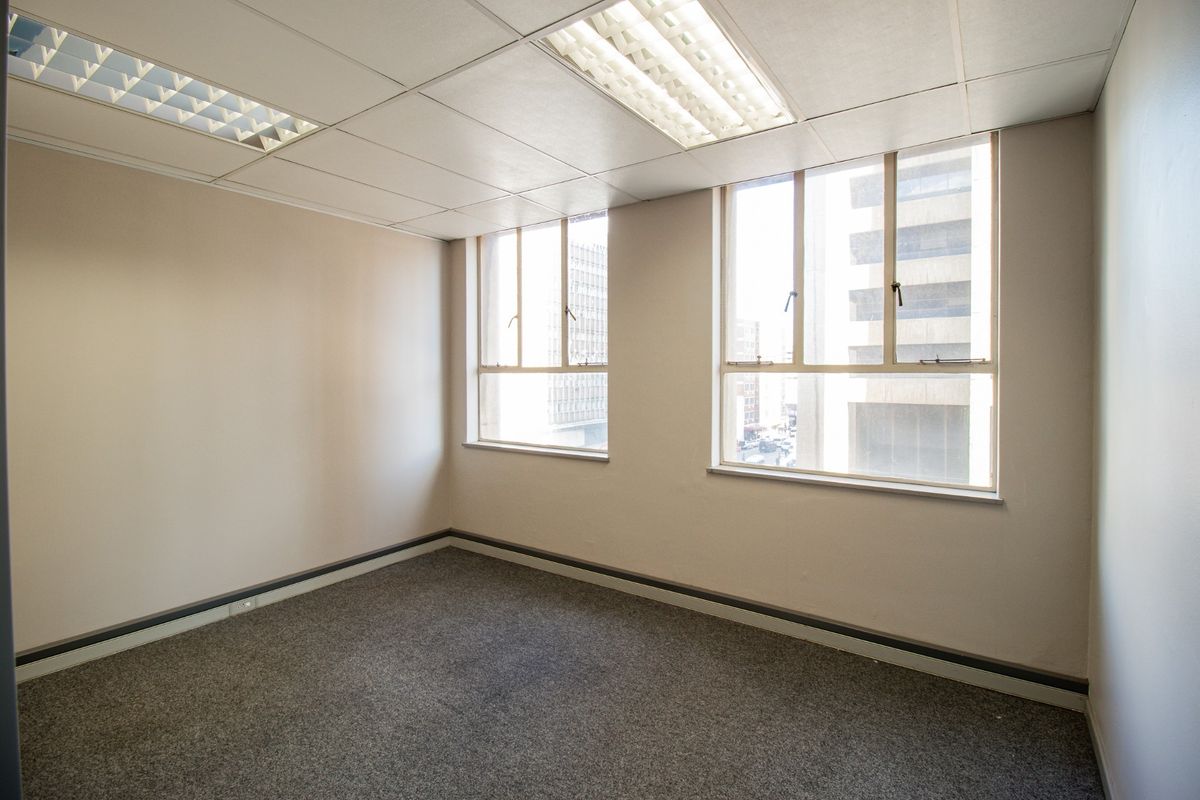 A vacant office space featuring neutral walls, grey carpeting, suspended ceiling tiles with recessed lighting, and large windows providing natural light.