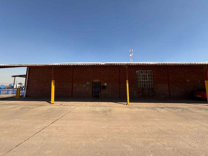Exterior view of an industrial warehouse building featuring a brick facade, metal roofing, and a large concrete loading or parking apron.