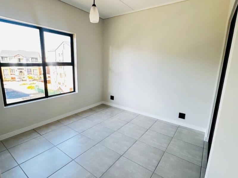A bright, empty bedroom featuring neutral tiled flooring, a modern pendant light, and a large window with views of the surrounding complex.
