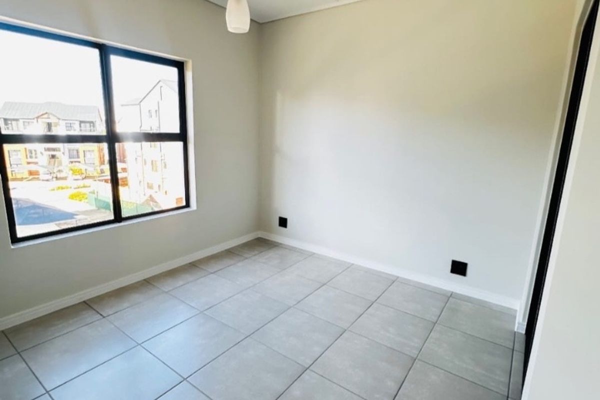 A bright, empty bedroom featuring neutral tiled flooring, a modern pendant light, and a large window with views of the surrounding complex.