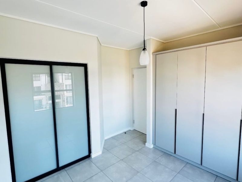 A bright, modern bedroom featuring neutral tiled flooring, ample built-in cupboard storage, and a sleek sliding glass door.