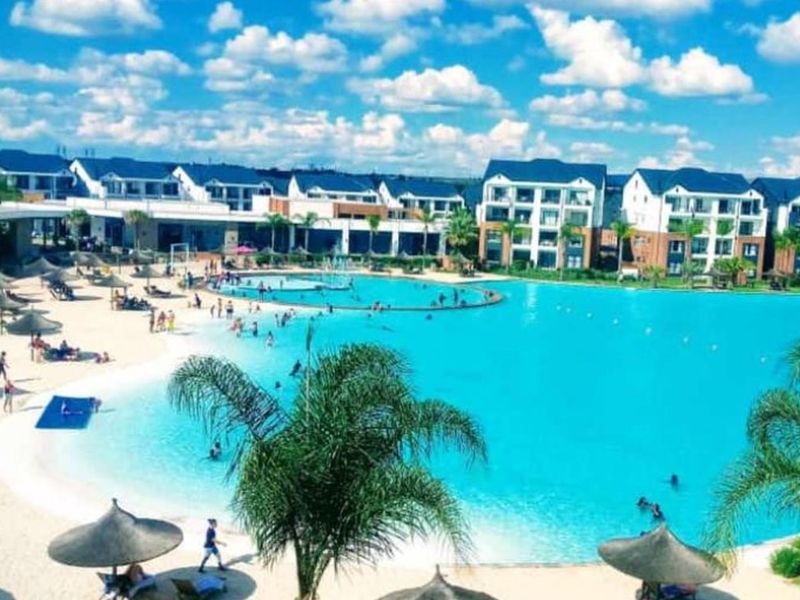 A large, resort-style swimming pool with a sandy beach area, surrounded by modern apartment buildings in a well-maintained residential complex.